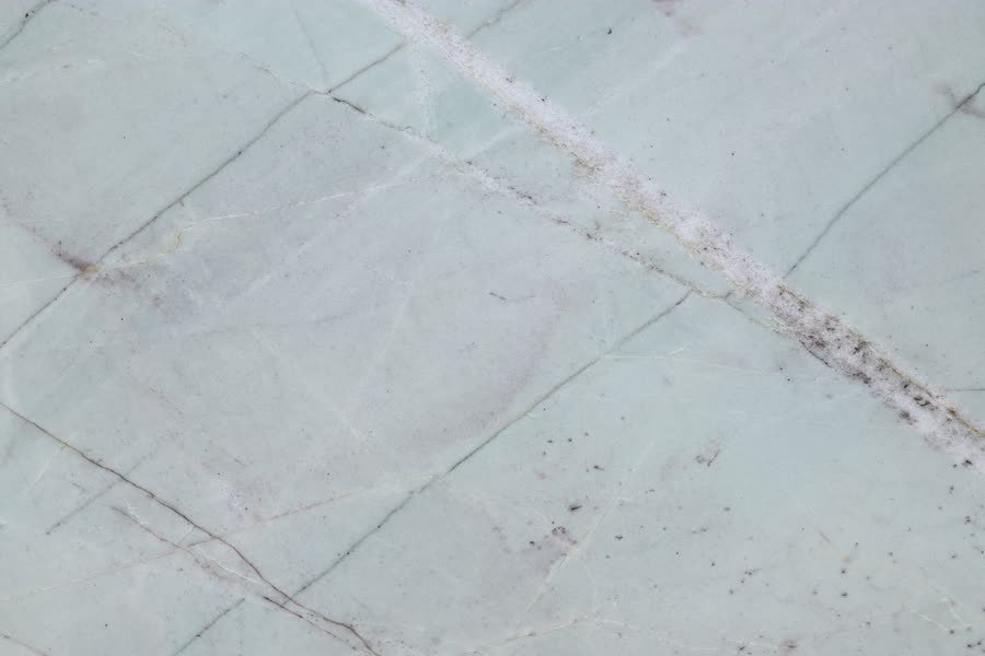 Close-up of a polished pale green-white stone surface, crossed by a broad diagonal seam of white crystalline mineral and finer rust and grey fissures.