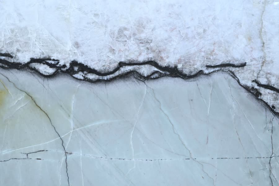 Macro view of a stone surface split horizontally: a white crystalline upper band, a jagged black vein running left to right, and a smooth pale green lower field crossed by fine grey and rust hairlines.