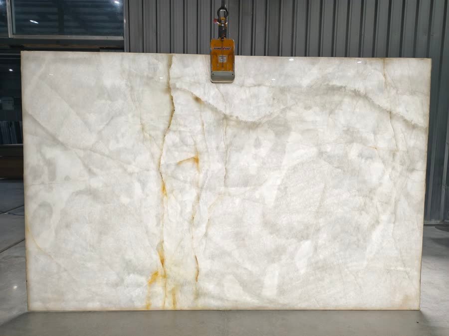 A large near-white translucent slab hung from an overhead lifting clamp in a warehouse, its milky crystalline surface crossed by soft grey shading and thin rust-gold veins running down the left of centre.