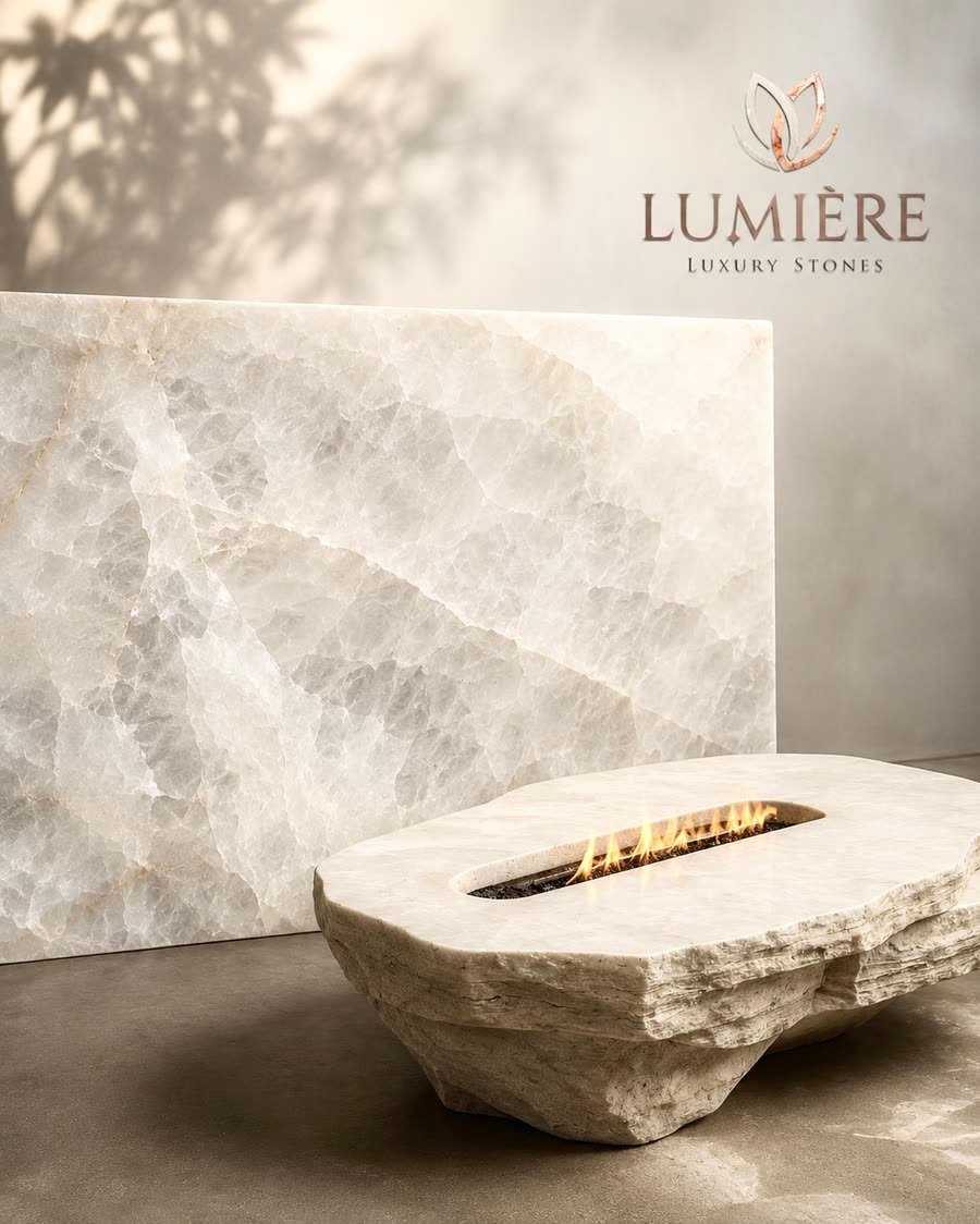 A cream-white crystalline stone panel leaning against a plaster wall dappled with palm-leaf shadow, with a carved stone fire bowl holding a low flame in front of it on a concrete floor, and the Lumiere Luxury Stones logo in the top right corner.