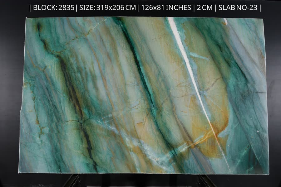 A full Mocamblu stone slab photographed upright on a display rack, its surface flowing with aqua, teal and gold veining and a bright white vein running top to bottom, with a printed specification strip along the top edge.