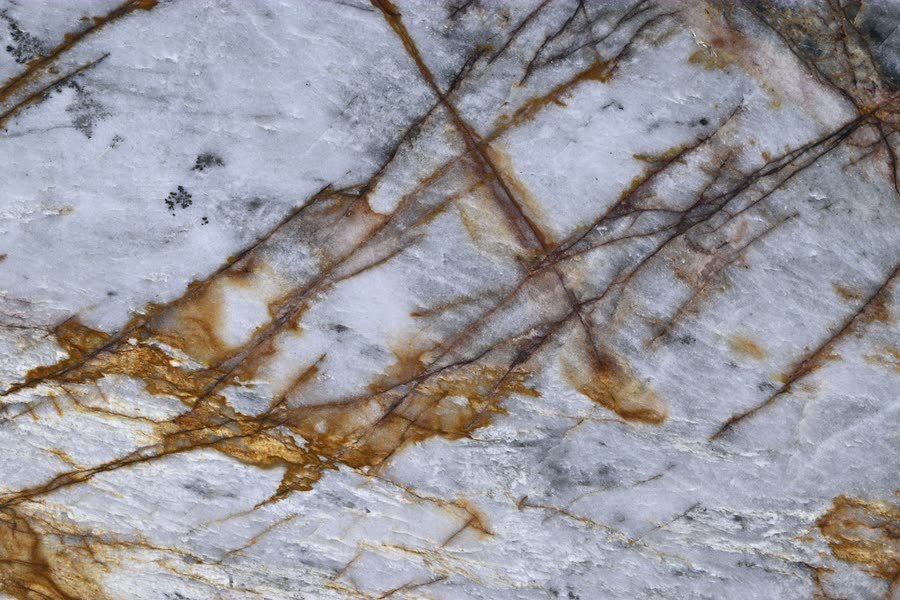 Macro close-up of a polished Nebula Crystal stone surface, translucent white and pale grey crossed by fine rust-gold and dark brown veins running diagonally, with a few small dark mineral flecks.