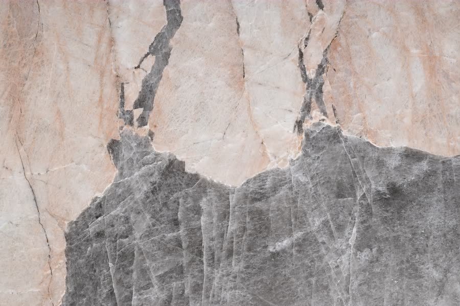 Extreme close-up of a polished stone surface where a pale pink and cream upper field meets a darker grey crystalline field along a jagged, mountain-like boundary, with fine dark fissures branching upward.