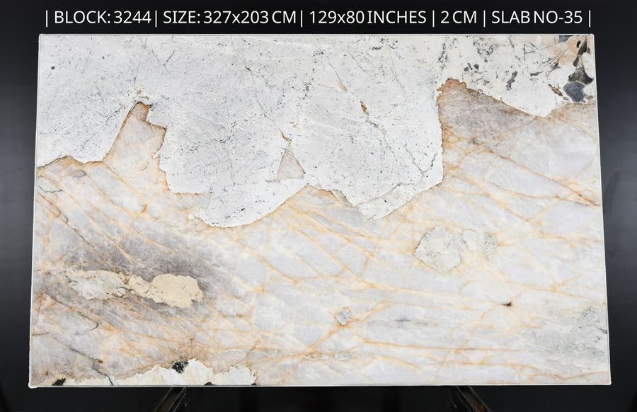 A full natural stone slab standing upright on a metal easel against a dark studio wall, its upper third a chalky white crust with grey speckling and the lower two thirds translucent pale stone crossed by fine amber and gold veins. A line of printed specification text runs across the top of the image.