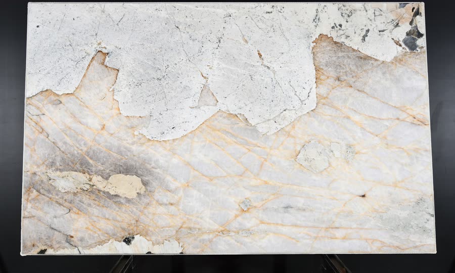 A full Patagonia slab on a metal easel against a dark studio wall: the upper third a chalky white crust with grey speckling, the lower two thirds translucent pale stone crossed by fine amber and gold veins.