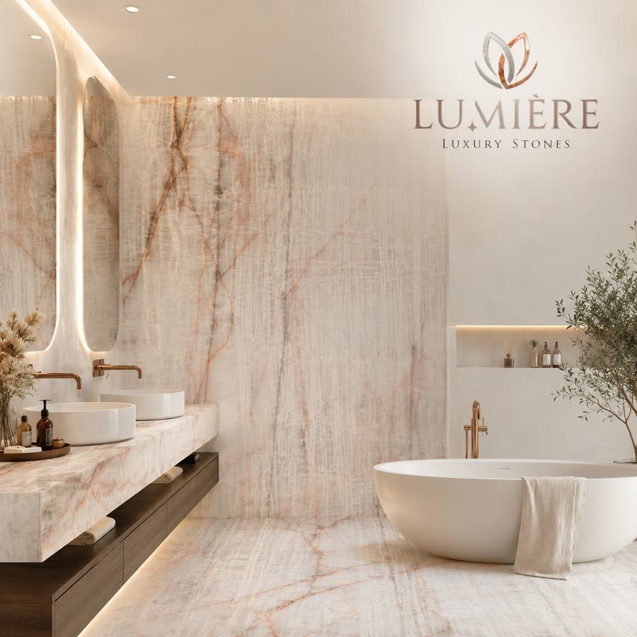 A bright bathroom rendered in warm neutrals: a floating stone-clad vanity with two round basins and brushed brass taps on the left, a tall backlit mirror, a large feature wall of pale stone with fine rust-orange veining, and a freestanding oval tub with a draped towel and an olive tree on the right. A lotus logo and wordmark sit in the upper right.
