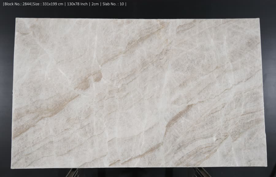 A large polished pale stone slab with soft beige and tan diagonal veining, stood upright on a metal A-frame against a black studio backdrop. A line of small white specification text runs across the top of the image.