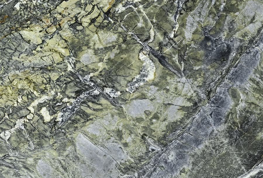 Close-up of a polished stone surface in mottled olive green, grey and cream, cut through by dark angular veins and pale crystalline seams.