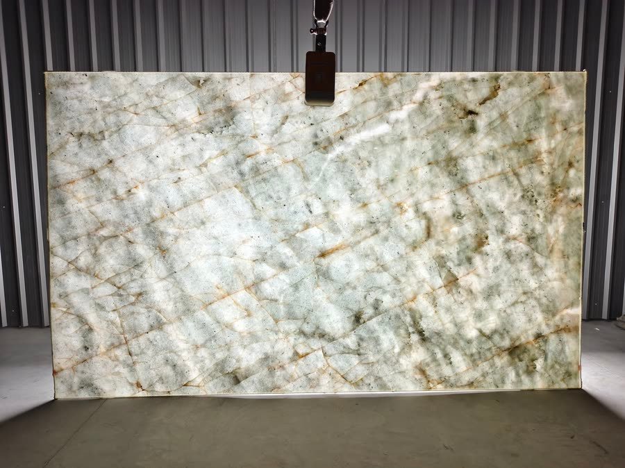 A large polished pale grey-green stone slab with white crystalline patches and rust-coloured seams, suspended upright by a lifting clamp against a corrugated metal wall on a concrete warehouse floor.