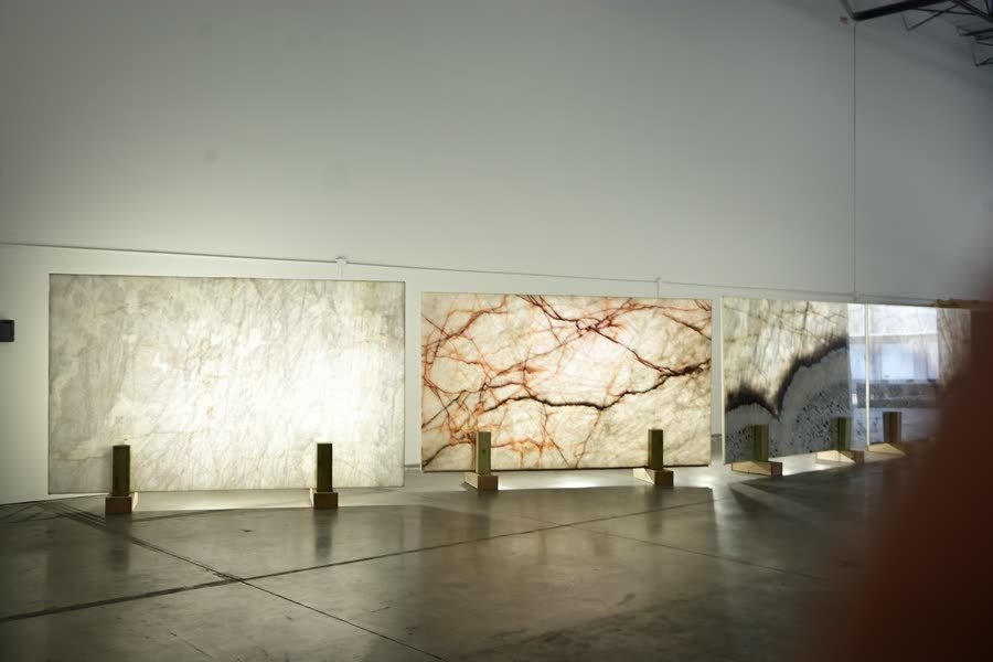 Three large backlit stone panels standing on brass plinths against a white wall in a gallery-like interior with a polished concrete floor. The left panel glows pale cream, the centre panel shows red and orange branching veins, and the right panel is banded in dark grey and white.