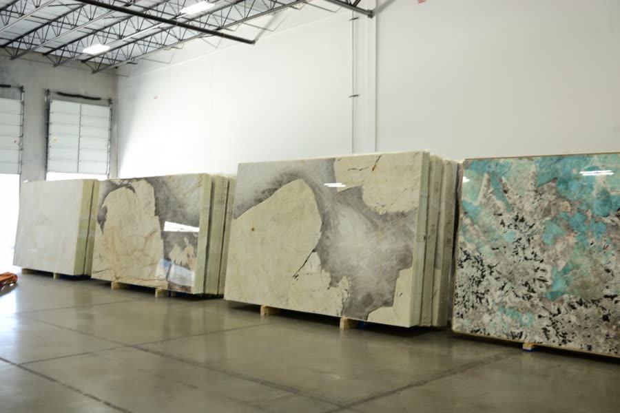 Several large stone slabs propped upright side by side in a warehouse: pale cream slabs at the left, a big white slab with sweeping grey and charcoal markings in the centre, and a turquoise and black speckled slab at the right.