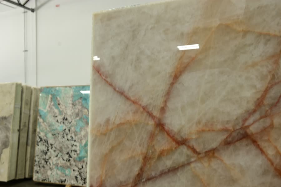 Close view of a large translucent white stone slab with fine rust-orange and red veins crossing its surface, lit by overhead strip lights; stacked cream slabs and a turquoise speckled slab stand behind it at the left.