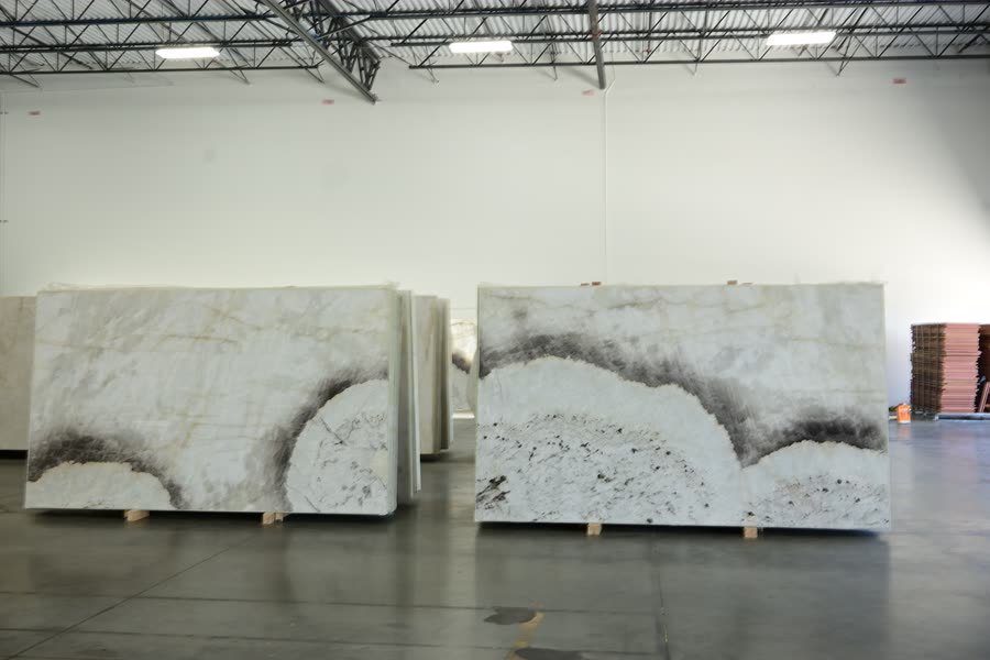 Two large stone slabs with matching white, grey and black cloud-like patterning standing side by side on wooden supports against a white warehouse wall, on a polished concrete floor.