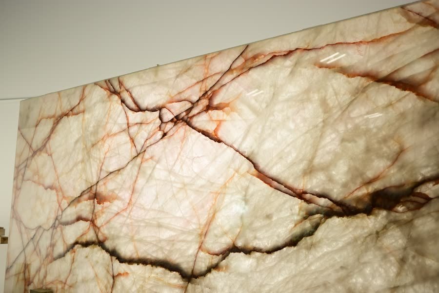 Close, angled view of a polished translucent white slab face crossed by rust-orange and dark brown veining, with small ceiling-light reflections on the surface.