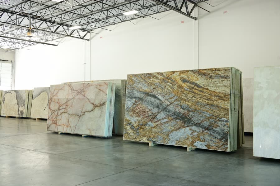Bundles of stone slabs standing upright on a polished concrete warehouse floor, the nearest with gold, grey and blue veining and the next with pink and red veining, lit by ceiling strips under a truss roof.