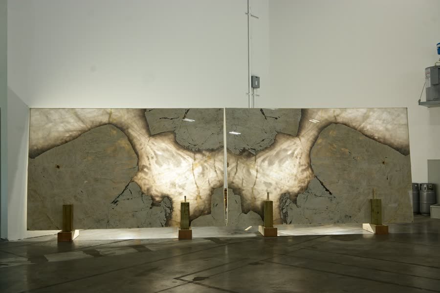 Two large stone slabs standing side by side on metal stands in a warehouse, mirrored so their pale, cloud-like veining forms a single symmetrical pattern across both panels against a white wall.