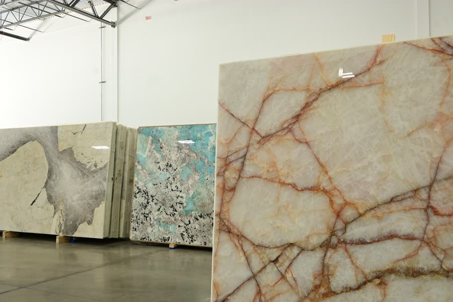 A large polished stone slab with a pale translucent background and branching orange-red veins standing upright in the foreground of a warehouse, with a grey-and-cream slab and a teal-green slab with dark speckling displayed on pallets behind it.