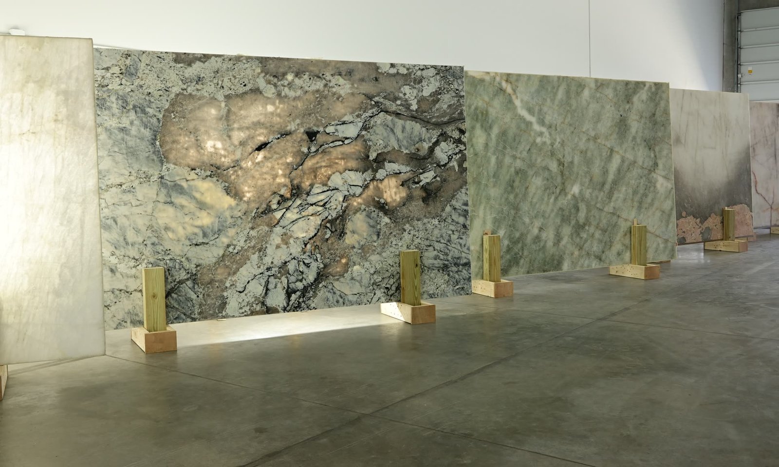 Full stone slabs standing on timber supports along a showroom wall, in grey, green and rose tones.