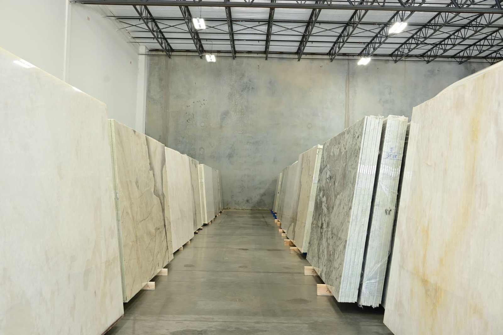 An aisle between two rows of stacked stone slabs on timber bearers, receding towards a concrete wall.